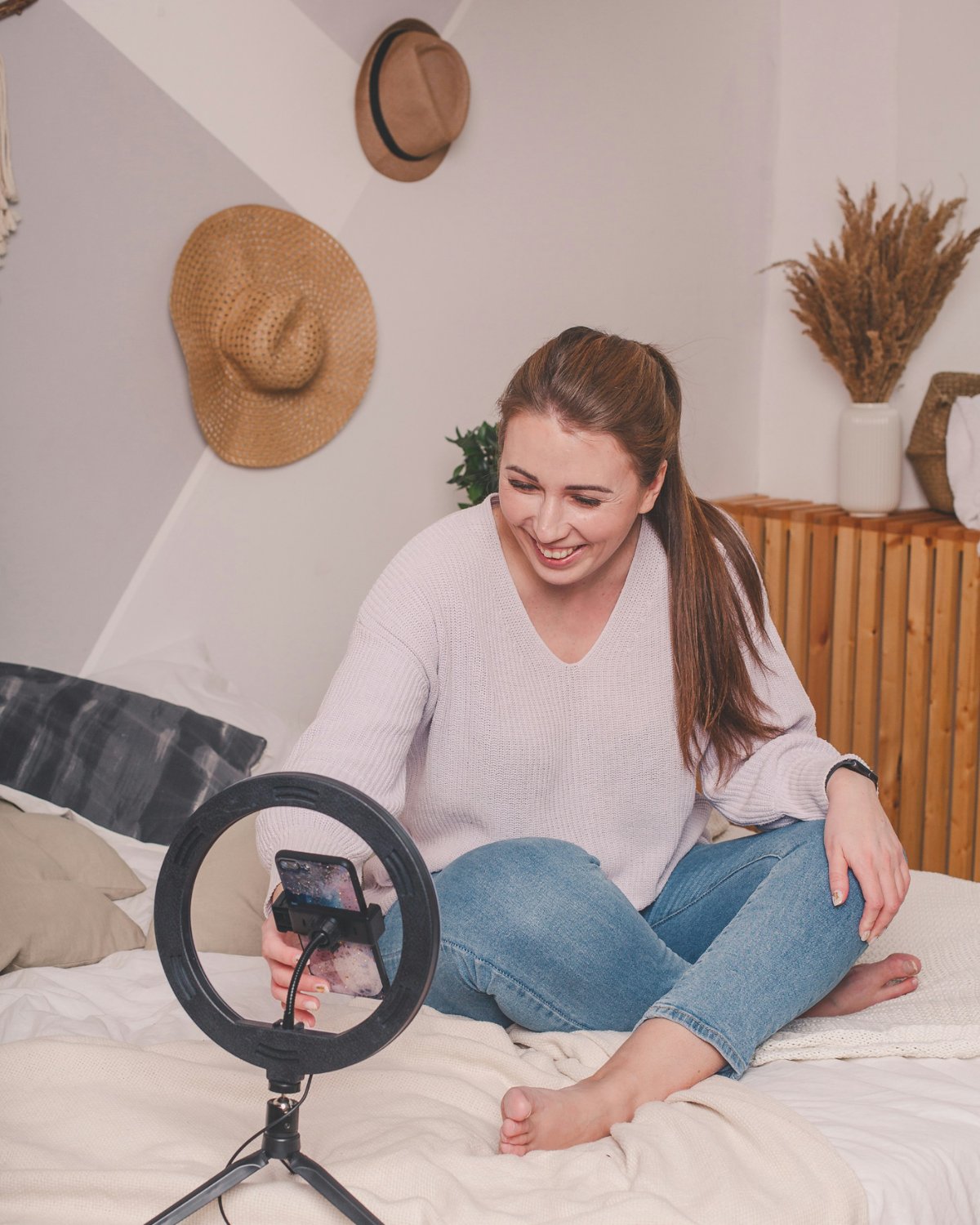 A smiling woman filming herself with a phone on a ring light, sitting on her bed